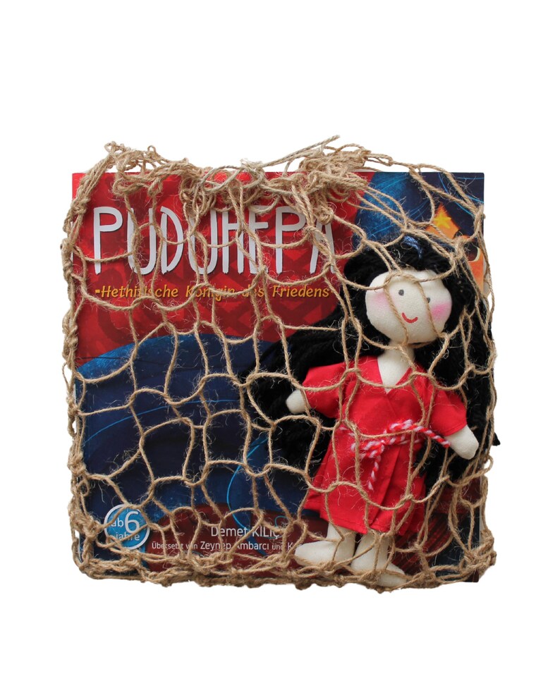 May include: A red and white doll with black hair is inside a brown burlap net bag. The bag is over a book with the title "PUDURHEPA" and the text "Hethitische Konigin des Friedens".
