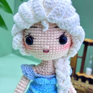 K&ouml;nnte beinhalten: Eine geh&auml;kelte Puppe mit einem wei&szlig;en Zopf, einem blauen Kleid und einem rosafarbenen Gesicht. Die Puppe hat schwarze Augen und einen l&auml;chelnden Mund.