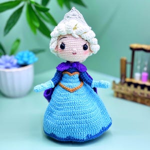 Könnte beinhalten: Handgefertigte Häkelpuppe einer Prinzessin in einem blauen Kleid mit goldenem Besatz, einem lila Umhang und einer weißen Krone. Die Puppe hat rosige Wangen, schwarze Augen und ein kleines Lächeln. Die Puppe ist etwa 20 cm groß.