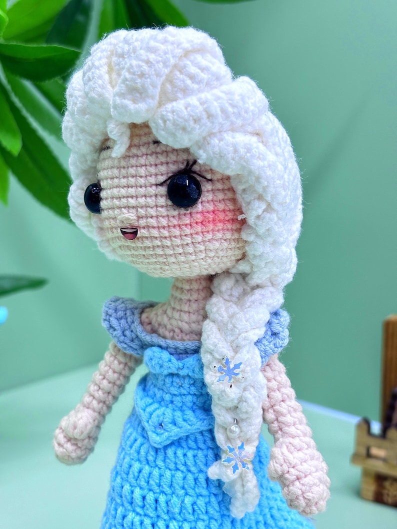 K&ouml;nnte beinhalten: Eine geh&auml;kelte Puppe mit einem wei&szlig;en Zopf und einem blauen Kleid. Die Puppe hat ein rosafarbenes Gesicht mit schwarzen Augen und einem L&auml;cheln. Der Zopf ist mit blauen Schneeflocken verziert.
