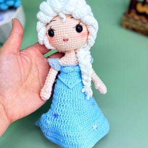 K&ouml;nnte beinhalten: Eine geh&auml;kelte Puppe mit einem wei&szlig;en Zopf, die ein hellblaues Kleid mit Schneeflocken-Stickerei tr&auml;gt. Die Puppe hat ein rosafarbenes Gesicht mit schwarzen Augen und einem l&auml;chelnden Mund.