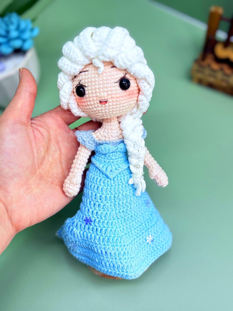 K&ouml;nnte beinhalten: Eine geh&auml;kelte Puppe mit einem wei&szlig;en Zopf, die ein hellblaues Kleid mit Schneeflocken-Stickerei tr&auml;gt. Die Puppe hat ein rosafarbenes Gesicht mit schwarzen Augen und einem l&auml;chelnden Mund.