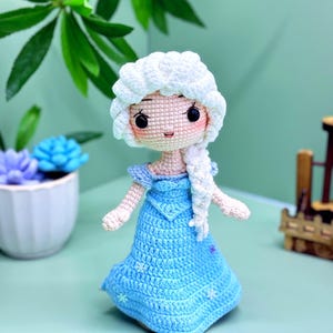 Könnte beinhalten: Eine gehäkelte Puppe mit einem weißen Zopf, die ein hellblaues Kleid mit weißen Schneeflocken-Akzenten trägt. Die Puppe hat eine weiße gehäkelte Krone und ein hellrosa Gesicht mit schwarzen Augen.