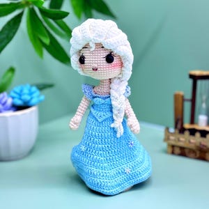 K&ouml;nnte beinhalten: Eine geh&auml;kelte Puppe mit einem wei&szlig;en Zopf, die ein hellblaues Kleid mit Schneeflocken-Akzenten tr&auml;gt. Die Puppe hat eine wei&szlig;e H&auml;kelm&uuml;tze und ein hellrosa Gesicht mit schwarzen Augen.