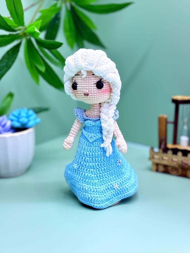 K&ouml;nnte beinhalten: Eine geh&auml;kelte Puppe mit einem wei&szlig;en Zopf, die ein hellblaues Kleid mit Schneeflocken-Akzenten tr&auml;gt. Die Puppe hat eine wei&szlig;e H&auml;kelm&uuml;tze und ein hellrosa Gesicht mit schwarzen Augen.