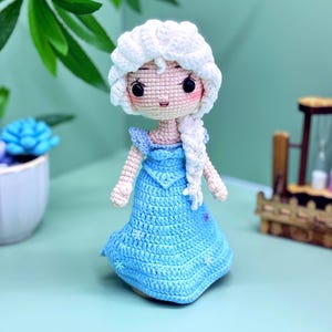 K&ouml;nnte beinhalten: Eine geh&auml;kelte Puppe mit einem wei&szlig;en Zopf und einem blauen Kleid. Die Puppe tr&auml;gt einen wei&szlig;en H&auml;kelhut und ein blaues Kleid mit wei&szlig;en Schneeflocken.