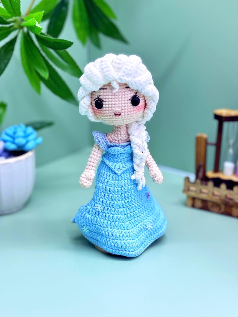 K&ouml;nnte beinhalten: Eine geh&auml;kelte Puppe mit einem wei&szlig;en Zopf und einem blauen Kleid. Die Puppe tr&auml;gt einen wei&szlig;en H&auml;kelhut und ein blaues Kleid mit wei&szlig;en Schneeflocken.