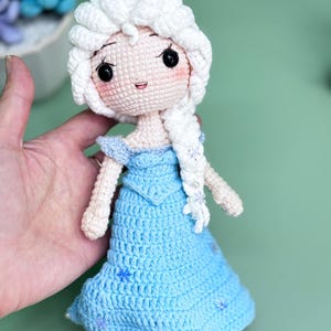 K&ouml;nnte beinhalten: Eine geh&auml;kelte Puppe mit einem wei&szlig;en Zopf, die ein blaues Kleid mit Schneeflocken-Details tr&auml;gt. Die Puppe hat eine wei&szlig;e H&auml;kelm&uuml;tze und einen s&uuml;&szlig;en Gesichtsausdruck.