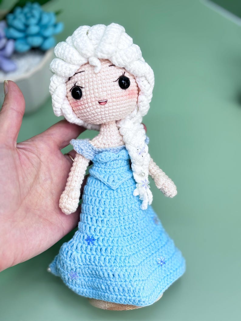 K&ouml;nnte beinhalten: Eine geh&auml;kelte Puppe mit einem wei&szlig;en Zopf, die ein blaues Kleid mit Schneeflocken-Details tr&auml;gt. Die Puppe hat eine wei&szlig;e H&auml;kelm&uuml;tze und einen s&uuml;&szlig;en Gesichtsausdruck.