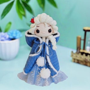 Peut inclure: Poupée au crochet faite à la main, vêtue d'un manteau à capuche bleu et blanc avec des bordures et des pompons blancs. La poupée a une coiffure tressée blanche, une fleur rouge et une robe bleu clair avec des détails de flocons de neige. La poupée mesure environ 15 cm.