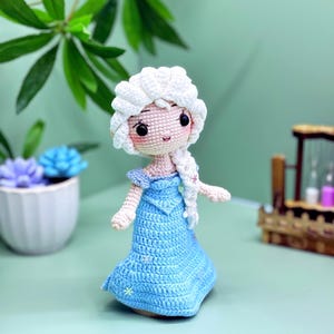 K&ouml;nnte beinhalten: Eine geh&auml;kelte Puppe mit einem wei&szlig;en Zopf, einem blauen Kleid und wei&szlig;en Schneeflocken-Akzenten. Die Puppe hat ein hellrosa Gesicht mit schwarzen Augen und einem hellrosa Mund.