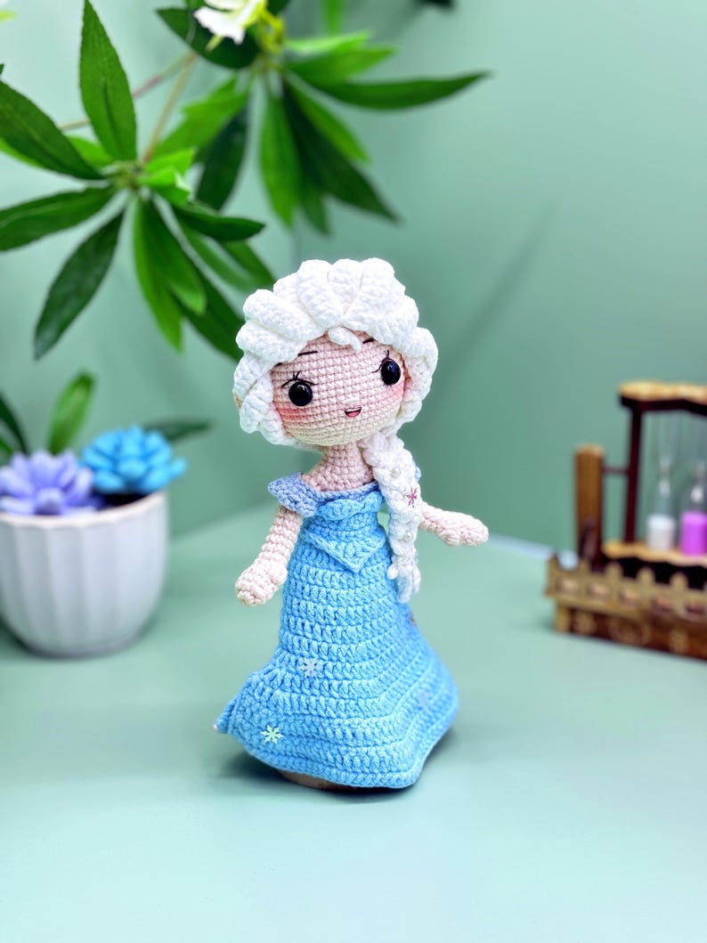 K&ouml;nnte beinhalten: Eine geh&auml;kelte Puppe mit einem wei&szlig;en Zopf, einem blauen Kleid und wei&szlig;en Schneeflocken-Akzenten. Die Puppe hat ein hellrosa Gesicht mit schwarzen Augen und einem hellrosa Mund.