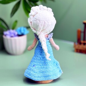 K&ouml;nnte beinhalten: Eine geh&auml;kelte Puppe mit langen wei&szlig;en Haaren in einem Zopf, die ein hellblaues Kleid tr&auml;gt. Die Puppe steht auf einer hellblauen Oberfl&auml;che.