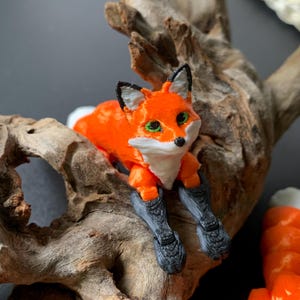 May include: A 3D-printed fox figurine in bright orange, white, and black, with green eyes. The fox is perched on a piece of driftwood, highlighting its detailed design and textured appearance. The figurine is a decorative item.