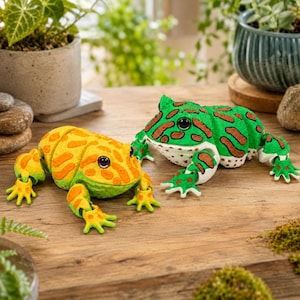 May include: Two articulated frog figurines with segmented bodies. One frog is bright yellow with orange spots and dark eyes, while the other is vibrant green with brown spots and dark eyes. Both feature detailed skin textures and splayed toes.