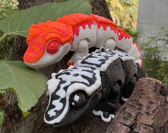 May include: Two 3D-printed gecko figurines. One is orange and white, the other is black and white. The geckos are perched on a tree branch with green leaves in the background. The figurines have detailed textures and realistic features.
