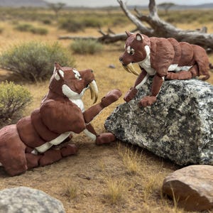Può includere: Due figurine di tigri dai denti a sciabola marroni e bianche in un ambiente naturale all'aperto. Uno è seduto su una roccia, l'altro è a terra, con le zampe alzate. Le figurine hanno grandi zanne ricurve.