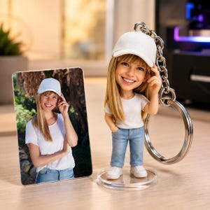 May include: A custom bobblehead keychain featuring a smiling person with long blonde hair, wearing a white baseball cap, a white t-shirt, and blue jeans, standing on a clear circular base, accompanied by a rectangular photo print of the same person.