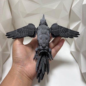 Puede incluir: Escultura detallada de un cuervo gris oscuro con alas extendidas. Las plumas y el cuerpo del ave están intrincadamente texturizados. La escultura se sostiene en una mano, mostrando su tamaño y detalle. La envergadura del cuervo es de aproximadamente 25 cm.