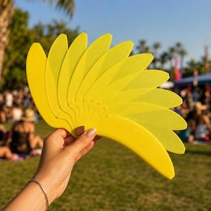 Puede incluir: Un abanico de mano de color amarillo brillante con múltiples láminas curvas en forma de pétalo que se despliegan desde un punto central. El abanico está construido con un material sólido de textura mate y se mantiene abierto.