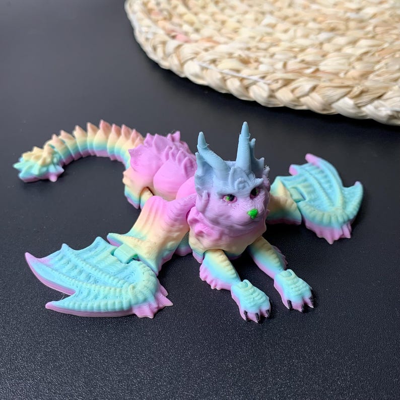 May include: A colorful, articulated dragon-cat figurine in pastel rainbow hues. The fantasy creature has a cat-like face with green accents, wings, and a long, segmented tail. The toy is made of plastic and is on a dark surface.