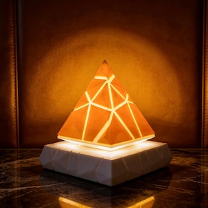 Könnte beinhalten: Eine geometrische pyramidenförmige Lampe mit warmem orangefarbenem Leuchten und einem weißen geometrischen Muster. Die Lampe steht auf einem weißen, facettierten Sockel, der von innen beleuchtet wird. Der Hintergrund ist eine dunkelbraune Lederstruktur.