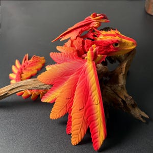 Peut inclure: Figurine de dragon articulé aux couleurs vives rouge, orange et jaune. Le dragon est posé sur un morceau de bois flotté, mettant en valeur les détails complexes de ses ailes et de ses plumes. La tête du dragon est jaune et rouge.