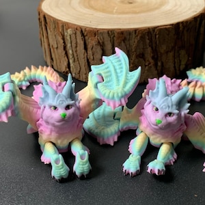 May include: Two pastel-colored dragon figurines with articulated limbs and wings. The dragons have pink, blue, yellow, and green hues. The figurines have a cat-like face with a green nose. The background includes a wooden slice.
