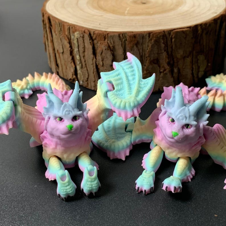 May include: Two pastel-colored dragon figurines with articulated limbs and wings. The dragons have pink, blue, yellow, and green hues. The figurines have a cat-like face with a green nose. The background includes a wooden slice.
