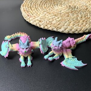 May include: Two articulated, pastel-colored dragon figures. Each dragon has a cat-like face with green nose, blue horns, and wings. The dragons are primarily pink, yellow, and blue, with a rainbow gradient effect. They are posed on a dark surface.