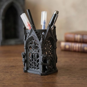 Peut inclure: Porte-stylo gothique noir avec des détails architecturaux complexes, notamment des fenêtres cintrées et des gargouilles. Le porte-stylo contient des stylos et des marqueurs. Le porte-stylo est en matériau sombre et posé sur une surface en bois.