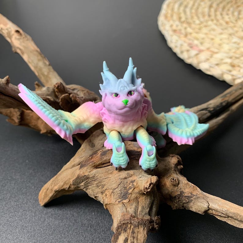 May include: A colorful fantasy creature figurine with a cat-like face, wings, and four legs. The figurine is primarily pastel pink, blue, and green. It is posed on a piece of driftwood against a dark background.