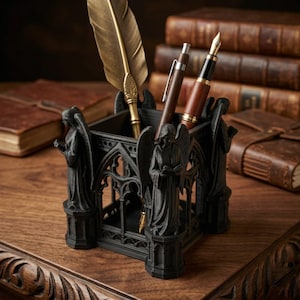 May include: A black pen holder with angel statues and gothic architectural details. The pen holder contains a gold feather quill and two pens. The background includes leather-bound books on a wooden surface.