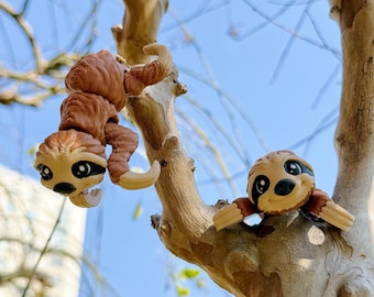 May include: Two brown sloth toys hanging from a tree branch against a blue sky. One sloth is hanging upside down, while the other is perched on the branch. The sloths have tan faces with black eyes and are made of plastic.