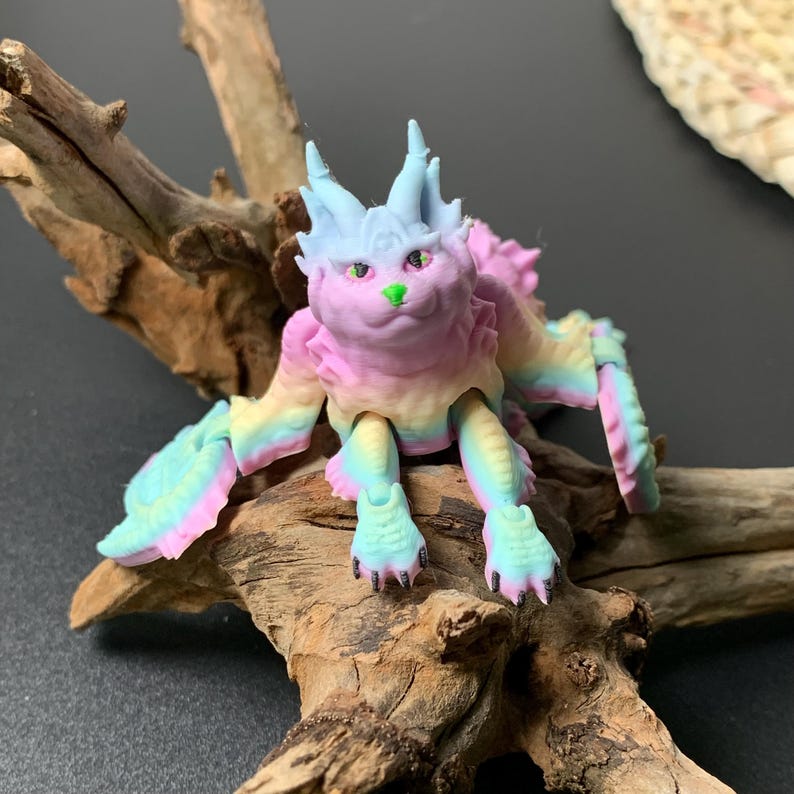 May include: A colorful fantasy creature figurine with a pastel rainbow color scheme. The figure has a cat-like face with green eyes and a pink nose. The wings and body are a mix of pink, blue, yellow, and green. The figurine is posed on a piece of driftwood.