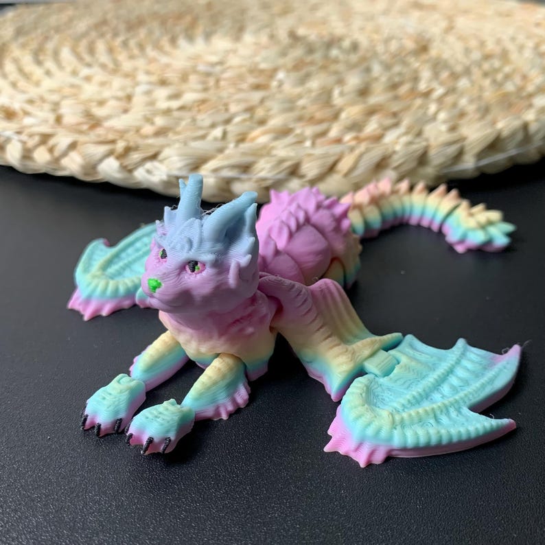 May include: A colorful, articulated dragon figurine in pastel shades of pink, blue, yellow, and green. The dragon has a cat-like face, wings, and a long, segmented tail. The figurine is lying on a dark surface.
