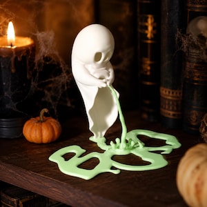 Peut inclure: Une figurine de fantôme blanche avec une tête arrondie et des yeux creux, tenant un mince filet de liquide vert clair qui s'écoule en une flaque plate et irrégulière sur la surface. Le fantôme a une finition mate lisse et une base drapée.