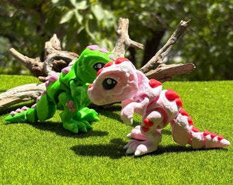 May include: Two toy dinosaurs, one green with pink accents and the other pink with red spots, are posed on a bed of green grass. The dinosaurs have large eyes and are in a playful stance, suggesting a children's toy.