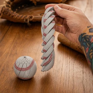 May include: A white baseball with red stitching, featuring a unique segmented design that allows it to be pulled apart into a long, flexible, spiraling chain of connected pieces, displayed alongside a complete, spherical baseball model.