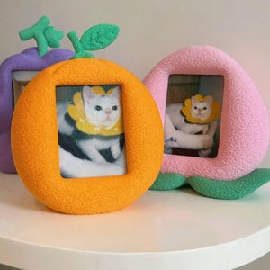 May include: Three whimsical photo frames in the shape of fruits: an orange, a peach, and a purple plum. Each frame holds a photo of a white cat wearing a yellow collar. The orange frame has a green stem and leaf.