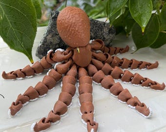 May include: A brown and white articulated octopus figurine. The octopus has a textured head and segmented tentacles. The tips of the tentacles are white. The figurine is resting on a white surface with green leaves in the background.