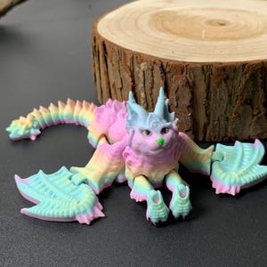 May include: A colorful, articulated dragon figurine in pastel shades of pink, blue, yellow, and green. The dragon has a cat-like face with horns and large, wing-like appendages. The tail is segmented, and the figure is posed on a dark surface.