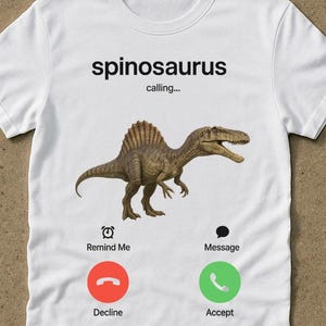 Könnte beinhalten: Weißes T-Shirt mit einem Spinosaurus-Dinosaurier-Design und dem Text "spinosaurus calling...". Das Design enthält Telefon-Interface-Symbole für "Remind Me", "Message", "Decline" und "Accept". Das Wort "white" befindet sich unten rechts.