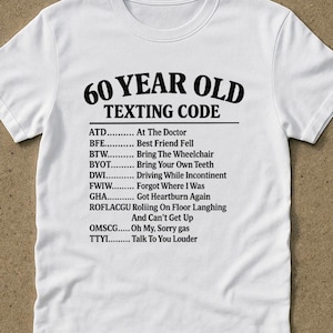 Può includere: Maglietta bianca con la scritta nera "60 YEAR OLD TEXTING CODE". La maglietta mostra un elenco di acronimi di messaggi di testo e i loro significati, come "ATD... At The Doctor" e "TTYI... Talk To You Louder".