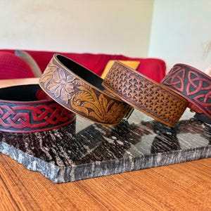 May include: Four leather bracelets with detailed patterns. One has a floral design, another a geometric pattern, and two with Celtic knotwork. The bracelets are in shades of brown and red, with black interiors. They are displayed on a grey stone surface.
