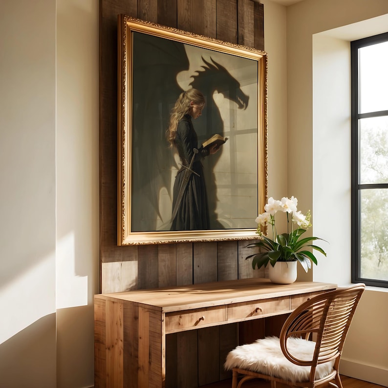 May include: A framed artwork depicts a woman in a dark dress reading a book, with a dragon shadow behind her. The artwork is in a gold frame and hangs above a wooden desk with a wicker chair.
