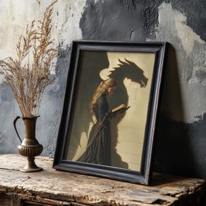 May include: A framed print featuring a woman in a black dress reading a book, with a dragon shadow on the wall. The print is in a black frame and rests on a wooden surface. A bronze vase with dried plants is to the left.