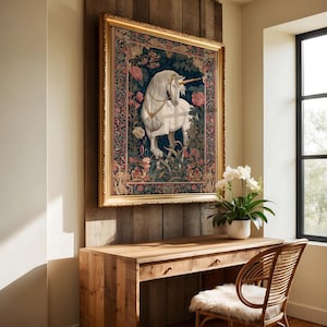 May include: A framed tapestry featuring a white unicorn with a golden horn, set against a dark blue background with floral and foliage details. The artwork is in a gold frame, mounted on a wooden wall above a wooden desk and chair.