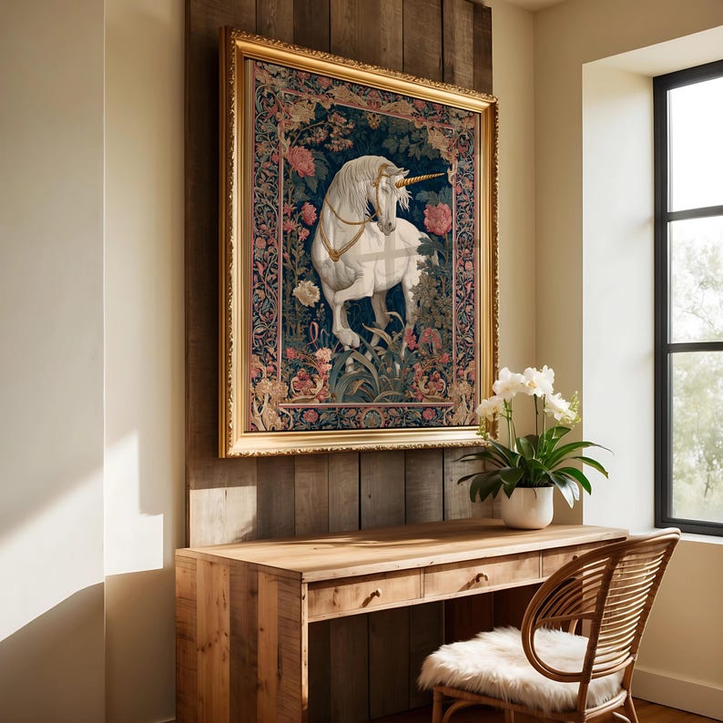 May include: A framed tapestry featuring a white unicorn with a golden horn, set against a dark blue background with floral and foliage details. The artwork is in a gold frame, mounted on a wooden wall above a wooden desk and chair.