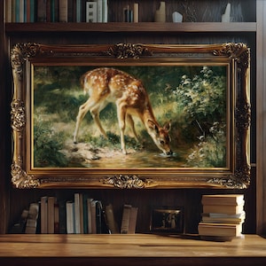 May include: A rectangular oil painting in an ornate, gilded gold frame depicting a spotted fawn drinking from a stream in a lush, green forest setting. The artwork features rich earth tones, deep greens, and soft light, capturing the animal in a natural, quiet moment within a dense woodland environment.